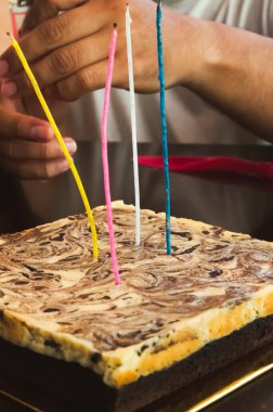 Koyu ahşap bir masada mermer çikolata ve cheesecake brownilerinin üst görüntüsü. Yetişkin ve çocuğun elleri pastaya mum koyuyor..