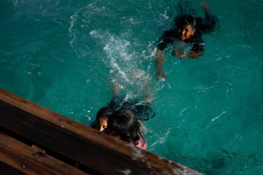 Çocuklar eğlence için kristal berrak mavi okyanusa atladıktan sonra bir iskelede merdivenleri tırmanırlar..