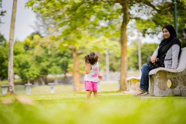 Annesi bankta otururken Asyalı çocuk parkta oynuyor.