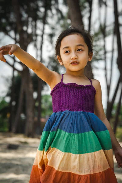 Renkli elbiseler giyen ve plaj ağaçlarının yanında duran güzel küçük kız..