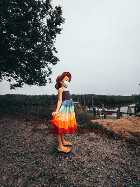 A little girl in a rainbow dress stands at the jetty. Summer day. Photo with selective focus and toning
