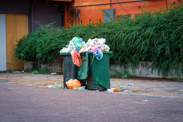 An overflowing trash can or garbage can with various plastic containers in an outdoor public park during weekend