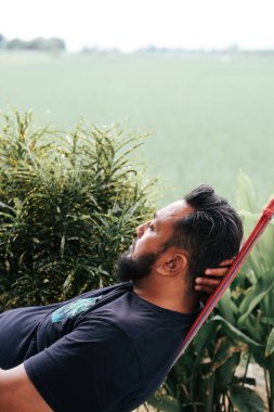 Asyalı sakallı bir adam hamakta oturmuş pirinç tarlalarına ve dağlara bakıyor. .