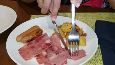 Kahvaltıda omlet, kızarmış patates, jambon ve beyaz tabakta sosis yiyorlar.