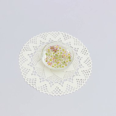 Altında dantel işlemeli su olan cam bir kasede renkli çiçekler. Açık mor kağıt arkaplan.