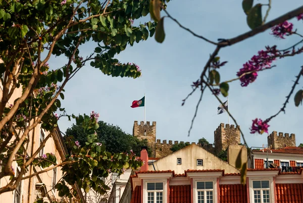 Rues de Lisbonne Drapeau portugais flottant dans le vent au-dessus de l'ancien fort avec des arbres en fleurs et des toits en tuiles rouges au premier plan - Lisbonne, Portugal — Photo