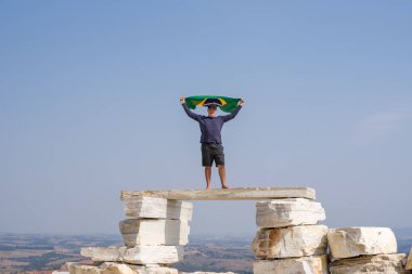 Brezilya Bayrağı ile Horizon 'a bakan, büyük düz kayaların üzerinde duran bir adamın uzak bir görüntüsü..