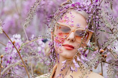 Güneş gözlüklü ve çiçek yapraklı güzel bir kadın çiçek açan salkımın dallarının etrafında.