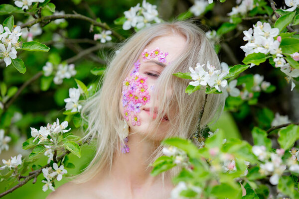 Beautiful woman with flowers petals on face around blooming apple tree
