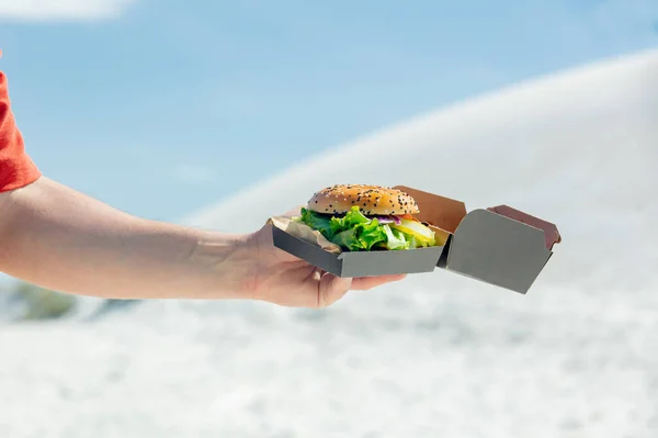 Man hold box with burger on a beach with sand on background