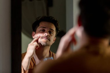 Adam banyoda aynanın yanındaki yüz derisine sıvı kremi sürüyor.