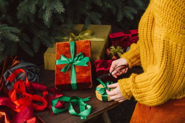 Woman in yellow sweater wrapping fabric eco friendly Christmas gift boxes on wooden table next to Christmas tree