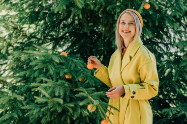 woman in yellow coat hangs oranges on the branches of the Christmas tree