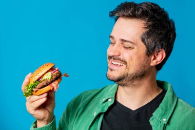 Mavi arka planda hamburgerli yeşil tişörtlü, gülümseyen beyaz adam.