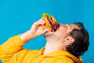 Sarı kapüşonlu mutlu beyaz adam mavi arka planda hamburger yiyor.