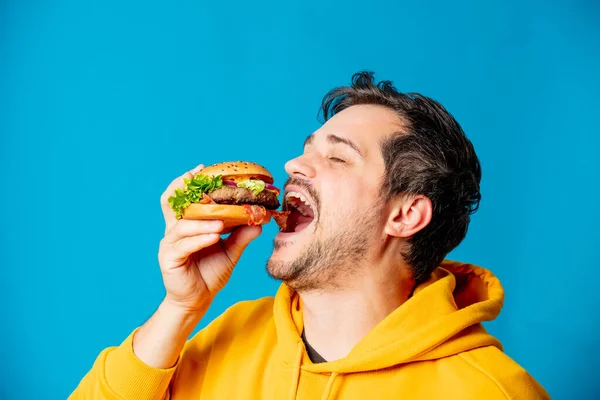 Gelukkig Wit Guy Geel Hoodie Eten Hamburger Blauwe Achtergrond — Stockfoto