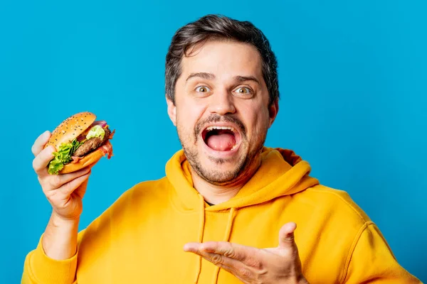 Happy White Guy Yellow Hoodie Eating Hamburger Blue Background — Stok Foto