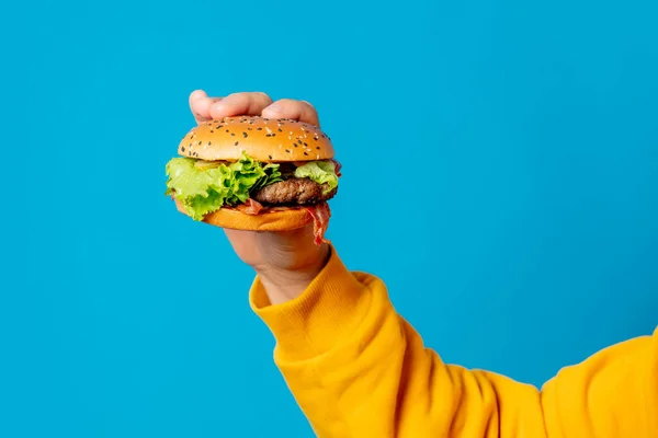 Mężczyzna Ręka Żółty Ubrania Trzymać Hamburger Niebieskim Tle — Zdjęcie stockowe