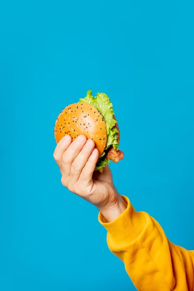 Male hand in yellow clothes hold hamburger on blue background