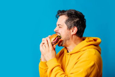  Sarı kapüşonlu beyaz adam mavi arka planda hamburger yiyor.