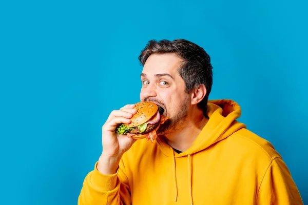 Wit Guy Geel Hoodie Eten Hamburger Blauwe Achtergrond — Stockfoto