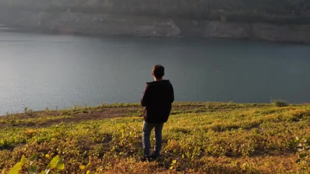 Observando el lago, el joven está observando el lago pensativamente a través de verdes praderas — Vídeo de stock