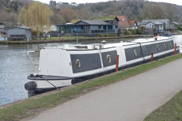 grey and white painted narrow boat moored at the river bank with houses and residences on the other side of the river