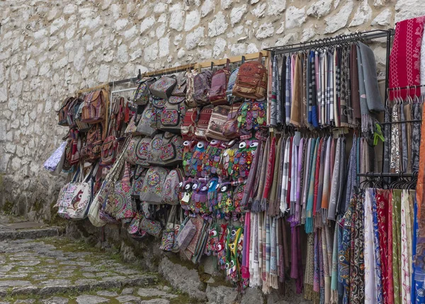 Şallar, battaniyeler ve el çantaları da dahil olmak üzere satılık parlak renkli mallar soluk renkli taştan bir duvara yaslanmış.