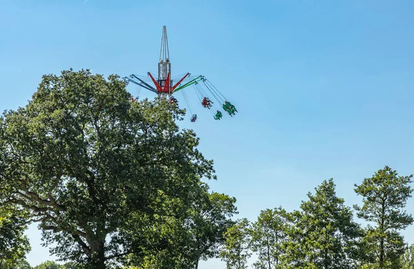 Fair ground ride visable behind the trees with people onboard sat in swinging seats