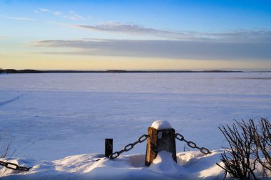 Zincirli bir kıyı taşı sütunu yüksek bir kar örtüsüne gömüldü. Merkezde ve arka planda, karla kaplı Volga Nehri ve karşı kıyıda, gün batımında ufukta kararıyor..