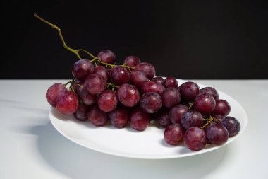 Tek bir üzüm kümesiyle hayat devam ediyor. Bir demet olgun sulu kırmızı üzüm beyaz bir tabakta zıt bir zemine uzanır..