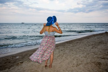 Genç bir kız akşamüstü deniz kıyısında elbisesiyle ve şapkasıyla, gün batımında, mavi gökyüzü, bulutlar, sakin, sörf, kum, boş kumsal, vahşi