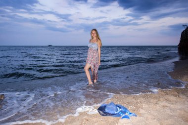 Genç bir kız akşamüstü deniz kıyısında elbisesiyle ve şapkasıyla, gün batımında, mavi gökyüzü, bulutlar, sakin, sörf, kum, boş kumsal, vahşi