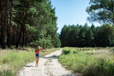 Genç bir kız yazın kozalaklı bir ormanın yakınındaki kumlu bir yolda yalınayak yürüyor. 