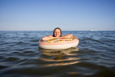 Şişme çemberdeki bir kız denizde yüzüyor, gülümsüyor.