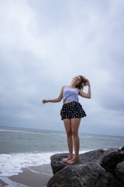 Beyaz bluzlu ve siyah şortlu genç bir kız denizin taşlarında duruyor, yaz mevsiminde.