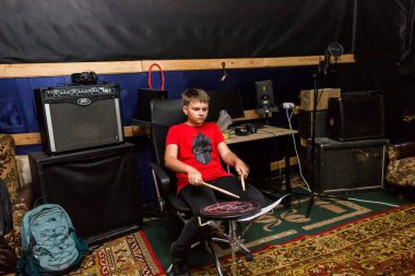 Genç bir davulcu davul çalar, sopayla çanlar çalar, özel bir sandalyede oturur.