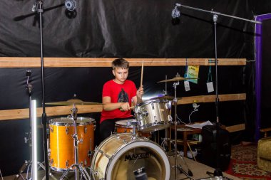 Genç bir davulcu davul çalar, sopayla çanlar çalar, özel bir sandalyede oturur.