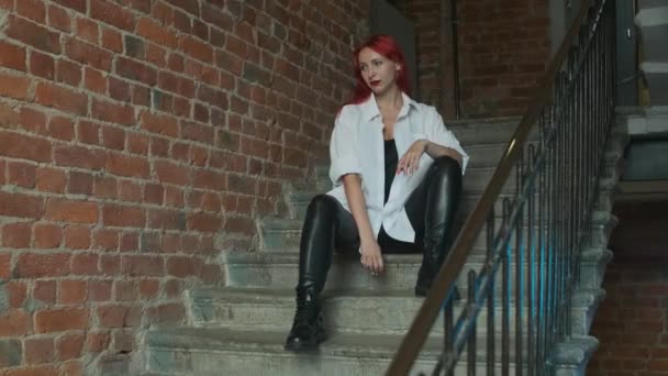 赤いレンガ造りの建物やフラットのブロックの階段に座ってまっすぐ見て彼女の鼻の中に革のズボン ベルト 黒と白の上とリングを身に着けている美しい赤髪の女の子 — ストック動画