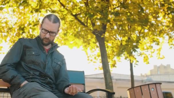 一个穿旧衣服戴眼镜的穷人坐在公园的长椅上 手里拿着硬币 一个破碎的人陷入贫穷 被解雇了 秋树背景 — 图库视频影像