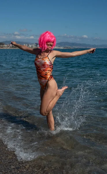 woman in swimming costume with slim athletic body getting her feet wet on the beach with positive attitude, pink hair, splashing water, arms raised, sea in background