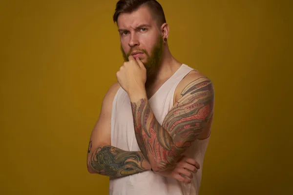 Brodaty mężczyzna w białej koszulce z tatuażami na ramionach, pozujący na poważny wygląd — Zdjęcie stockowe