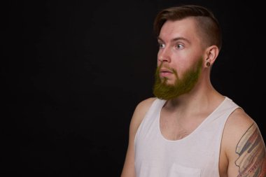 Kollarında dövmeleri olan beyaz tişörtlü şık sakallı bir adam.