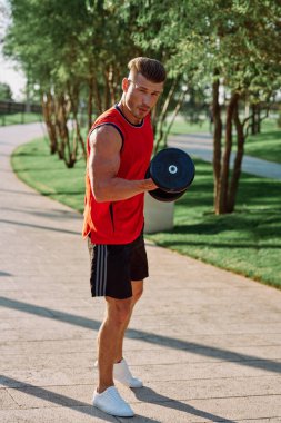 Parkın içinde elinde dambıllar olan atletik bir adam.