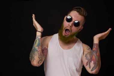 Kollarında dövme olan yakışıklı bir adam. Güneş gözlüğü takıyor. Geçmişi koyu renk.