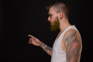 Kollarında dövmeleri olan beyaz tişörtlü, kabadayı pozu veren sakallı bir adam.