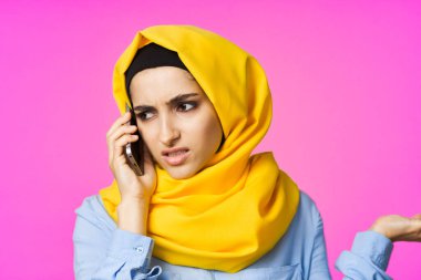 Sarı tesettürlü müslüman kadın telefonda pembe teknolojiyle konuşuyor.
