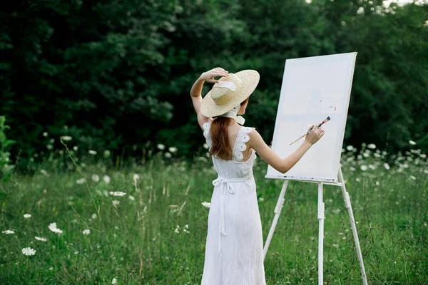 Eine Frau im weißen Kleid auf einem Feld mit Blumen malt ein Bild — Stockfoto