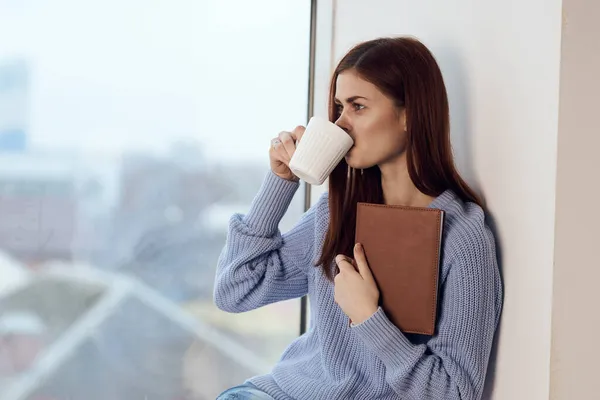 Mujer leyendo un libro cerca de la ventana con una taza de descanso bebida — Foto de Stock