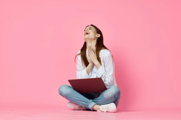 Mujer alegre con el ordenador portátil sentado en el suelo sobre un fondo rosa — Foto de Stock